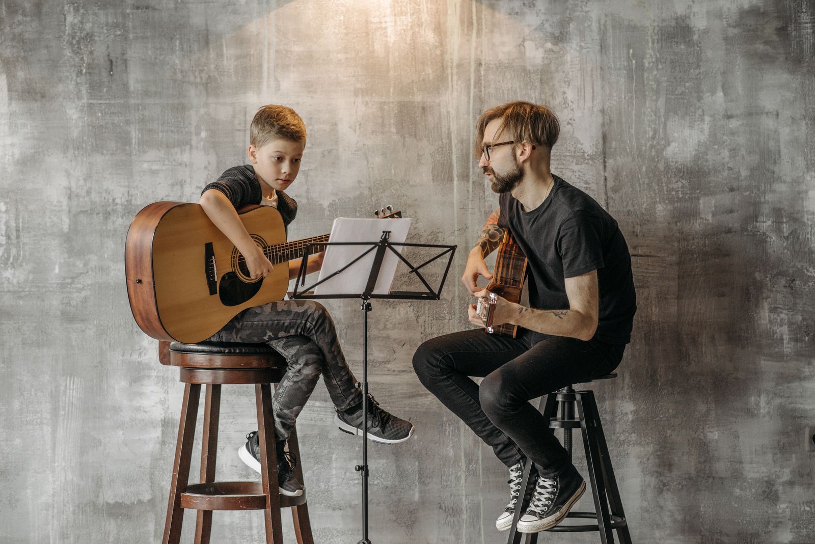 A teacher and a young boy sitting on stools with acoustic guitars and a music stand